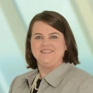 A woman with shoulder-length brown hair wearing a light gray blazer, smiling against a light green background.