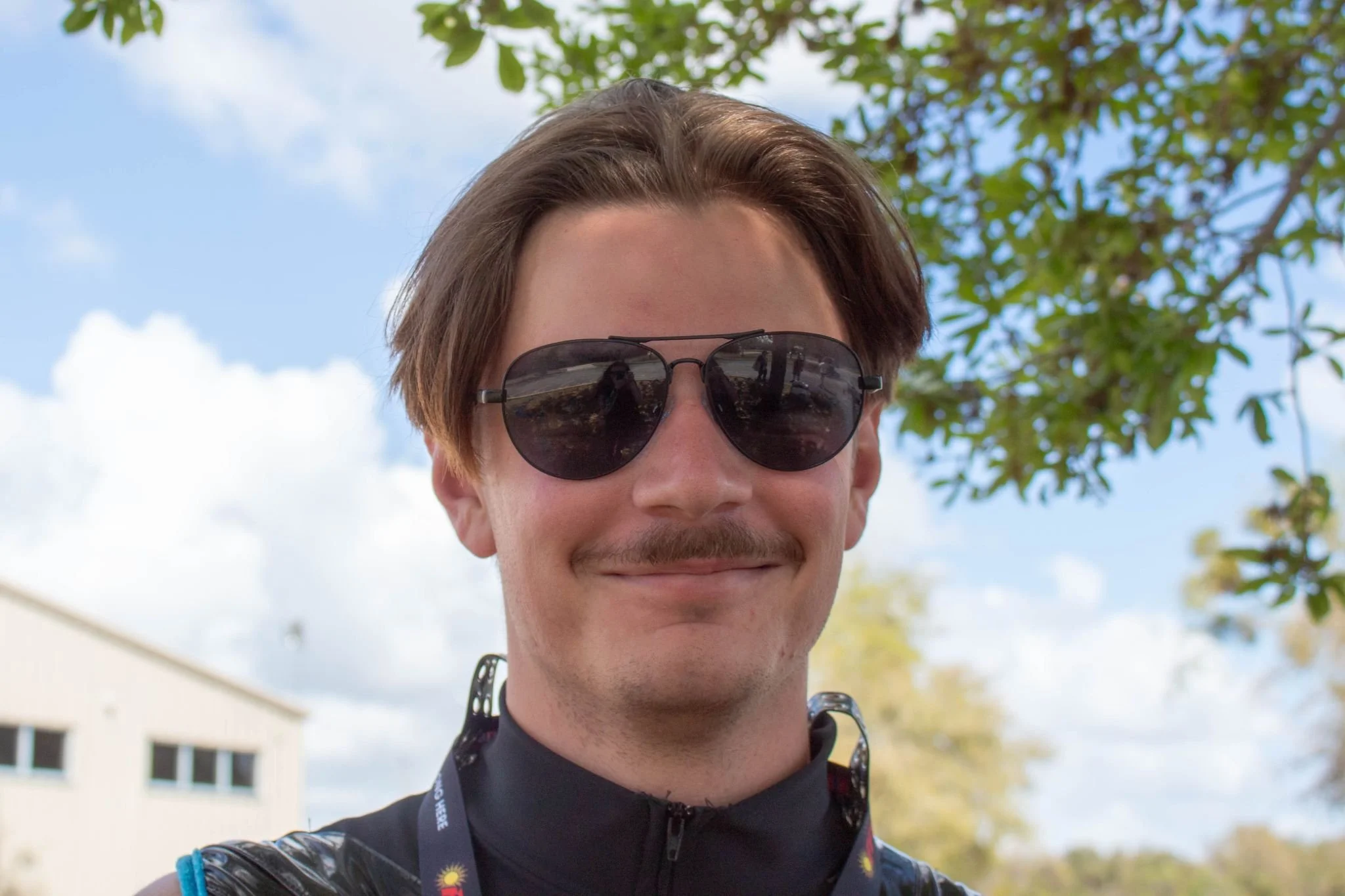 A man wearing sunglasses and a black jacket smiles outdoors with a tree and a blue sky in the background.