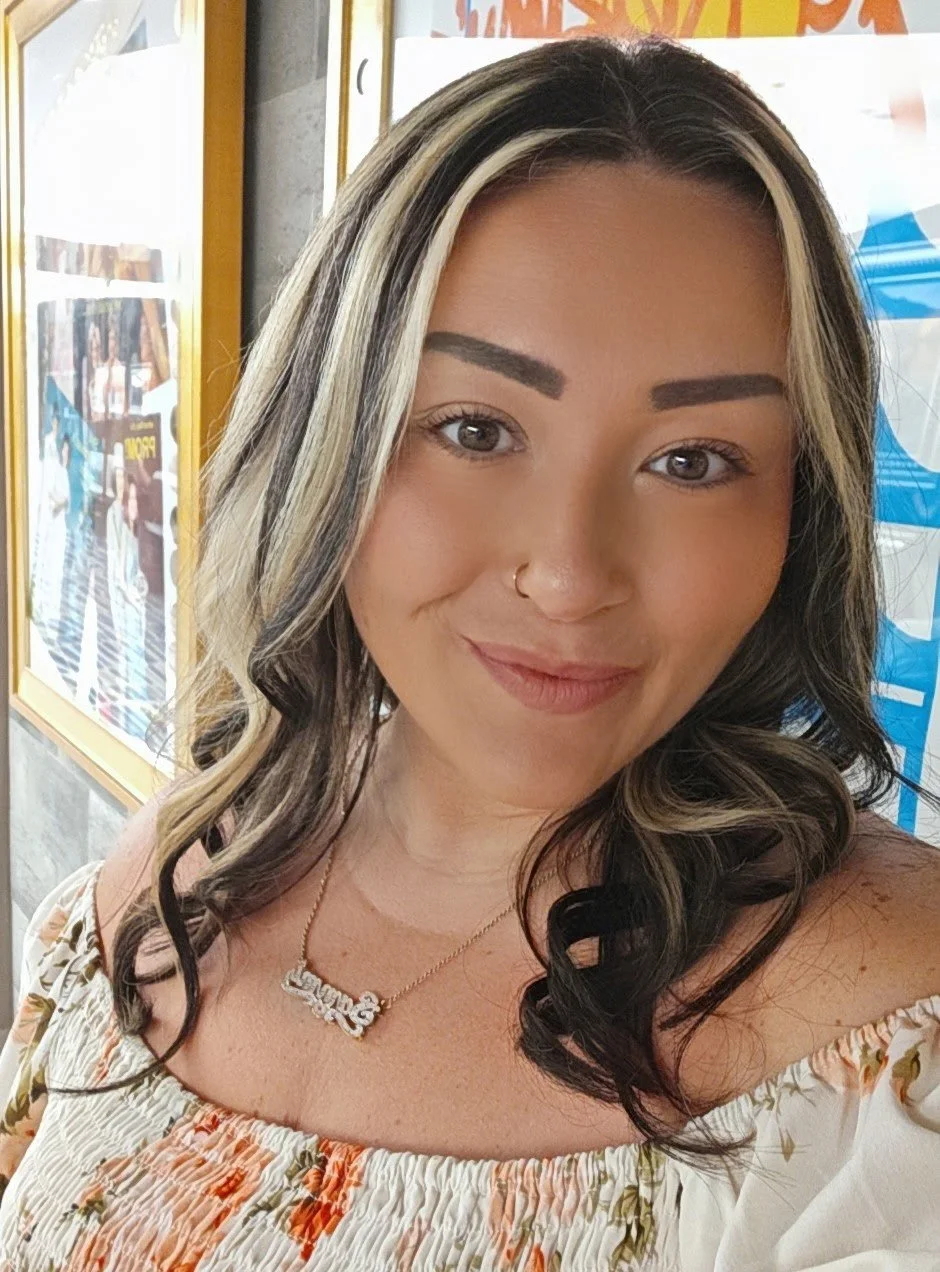 A young woman with light brown skin, dark hair with blonde highlights, and a nose ring, smiling for a selfie in an indoor setting with colorful posters in the background.