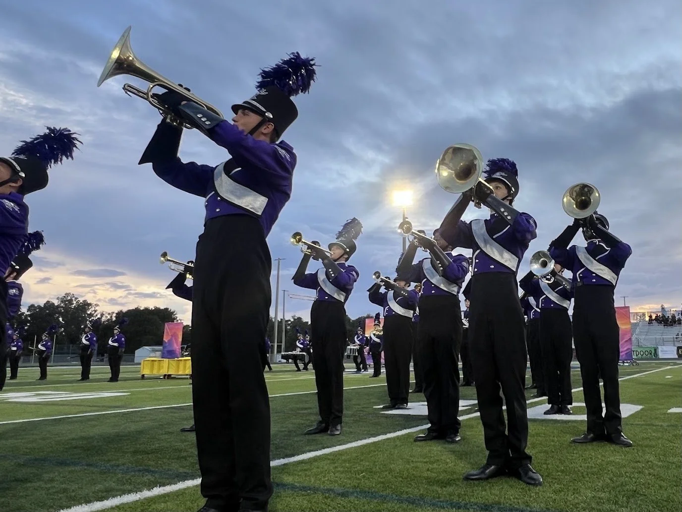 Marching band members dressed in purple and black marching band uniforms playing trumpets on a football field during sunset.