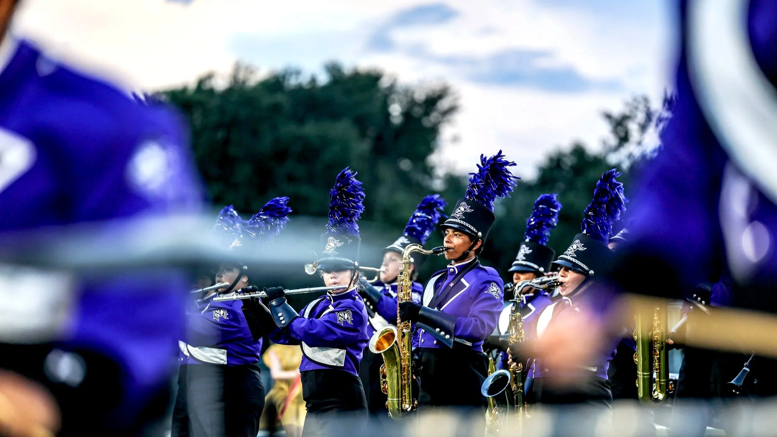 A marching band performing outdoors, members dressed in purple uniforms with black hats adorned with purple plumes, playing wind instruments during a parade or event.