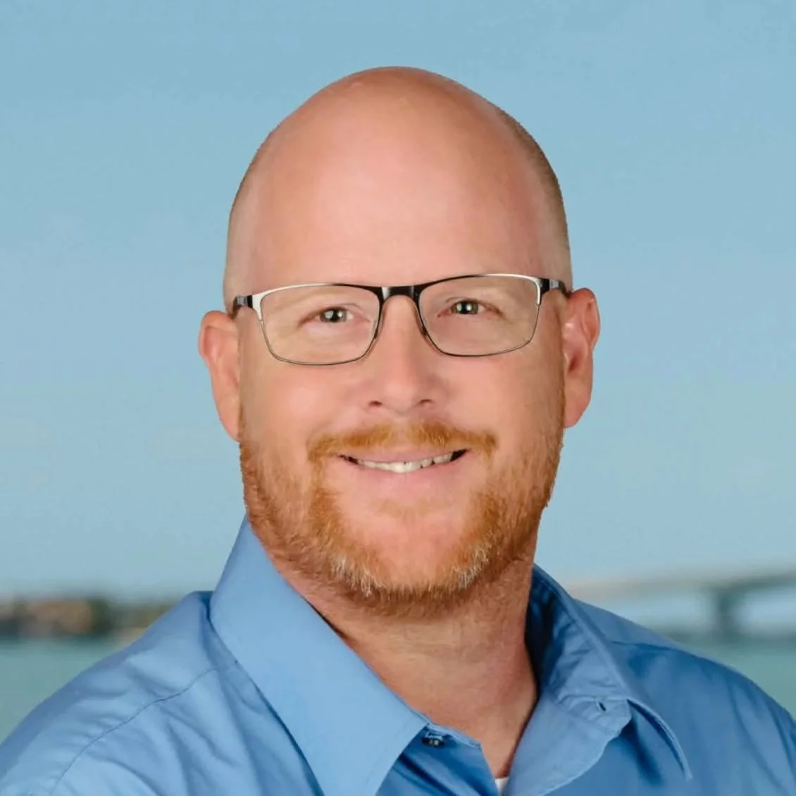 A smiling man with glasses, a beard, and a shaved head, wearing a blue shirt, against a blurred outdoor background.