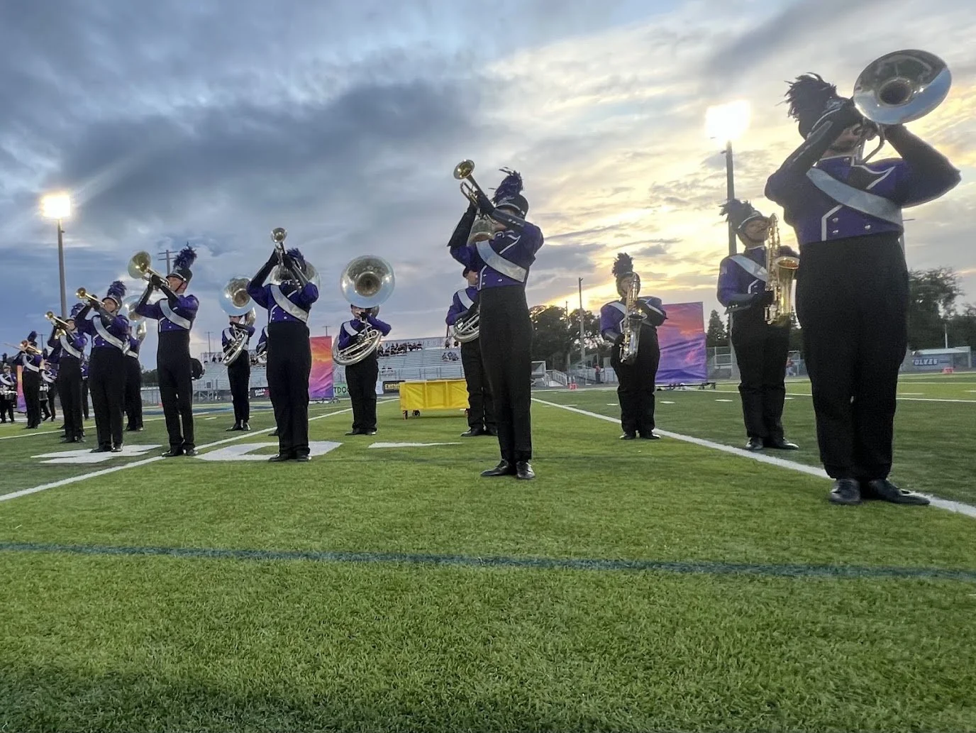Musicians in marching band uniform playing instruments on a football field during sunset.