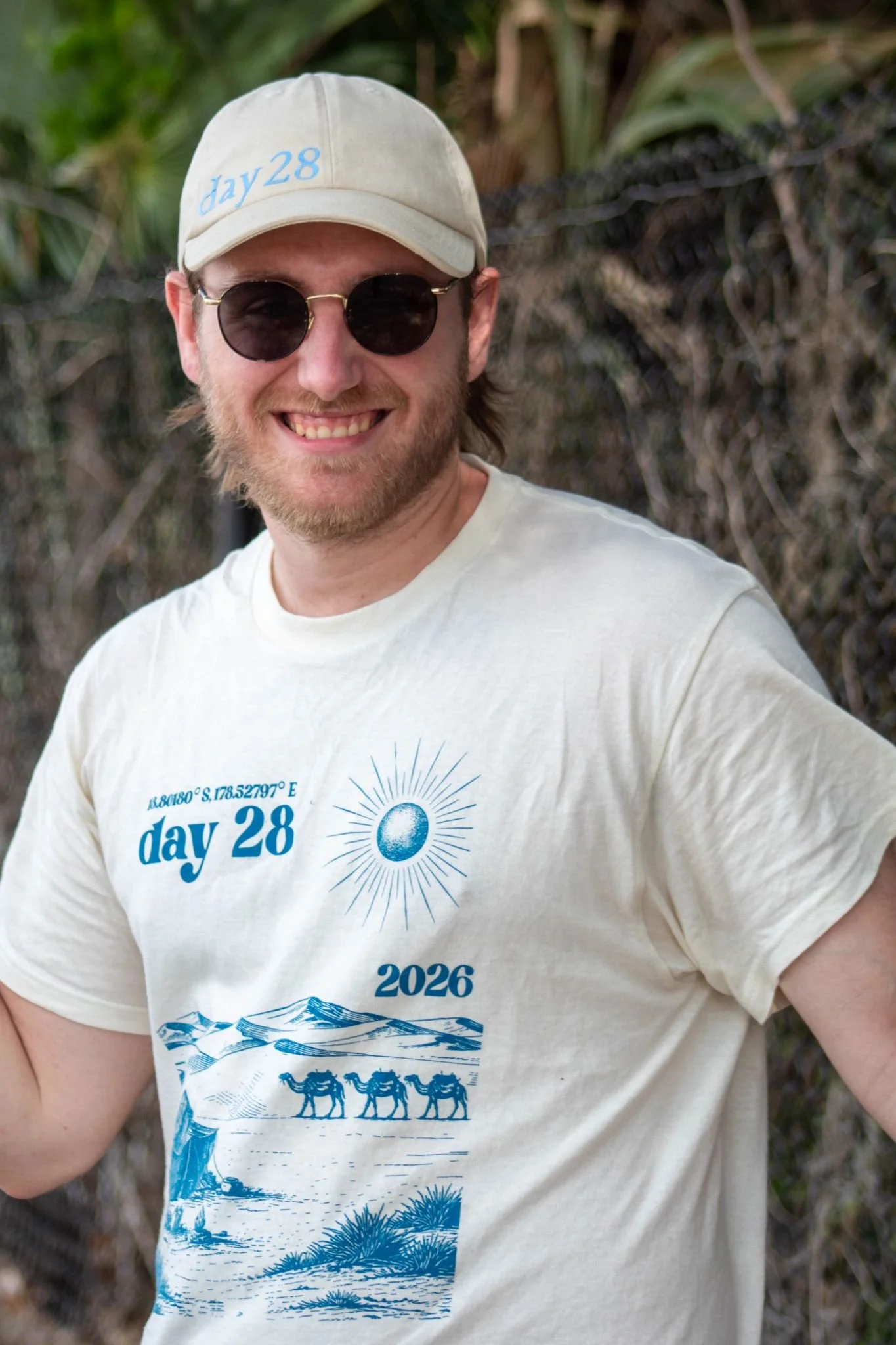 A man wearing a beige cap with 'day 28' embroidered on it, black sunglasses, and a white T-shirt with blue prints and text that reads 'day 28,' '2026,' and a mountain scene with camels. He is smiling and standing outdoors with a background of trees and bushes.