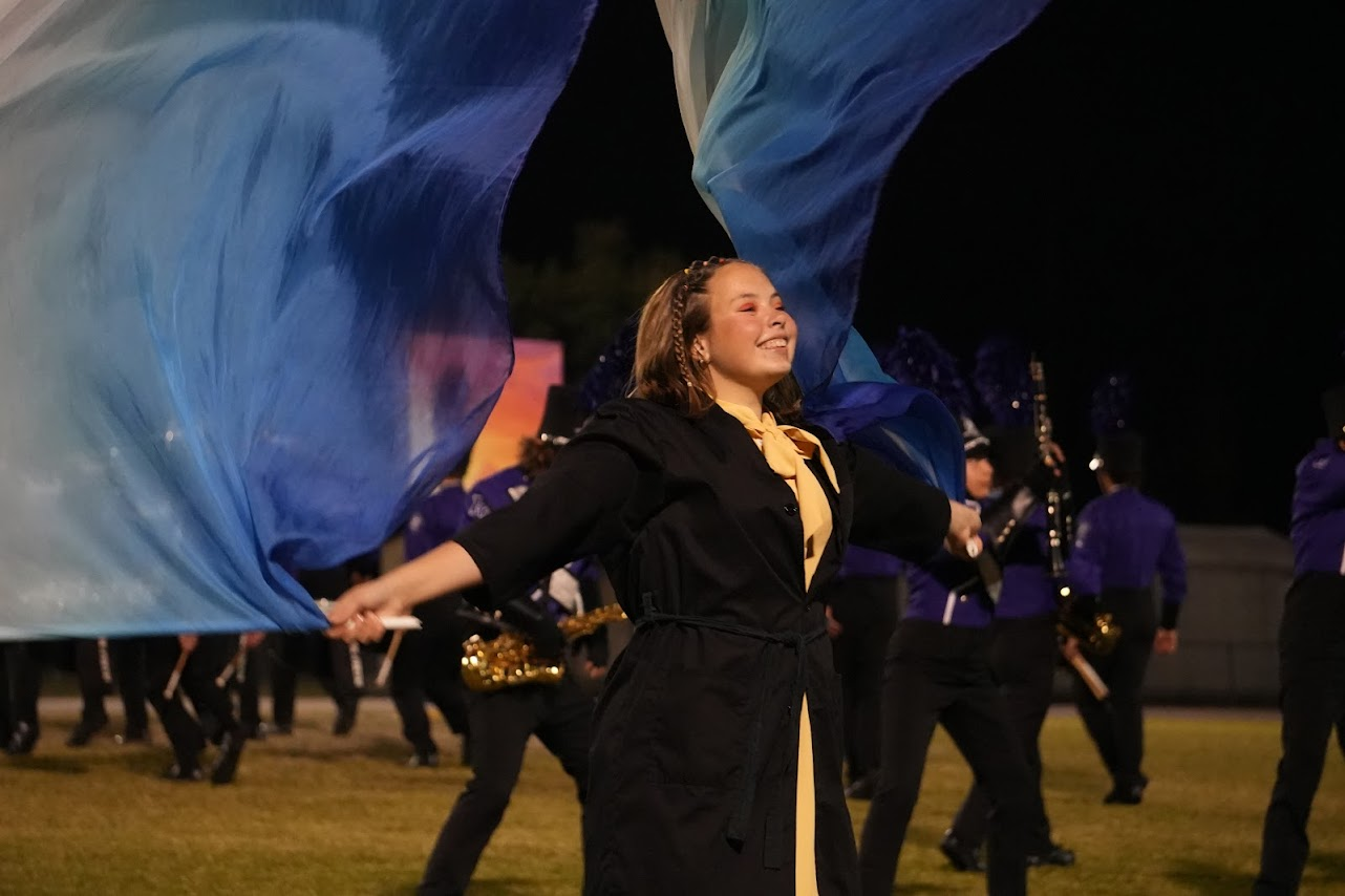 A young woman in a black jacket with a yellow scarf and braid, smiling as she twirls a large blue flag during a nighttime marching band performance, with band members playing instruments in the background.