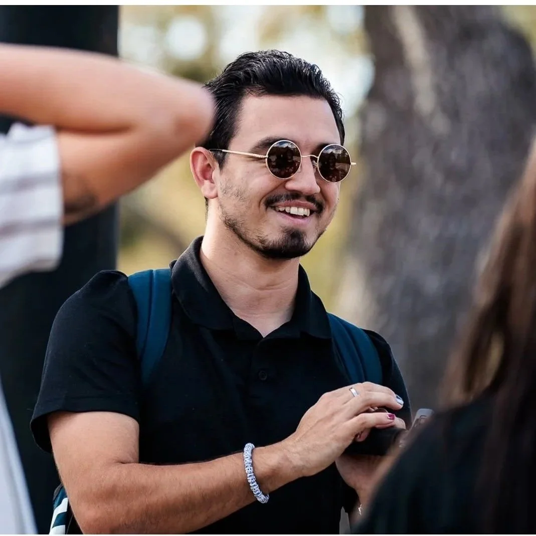 A man with dark hair, sunglasses, and light facial hair smiling outdoors, wearing a black polo shirt and holding a phone.