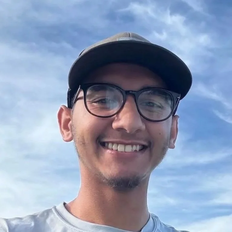Smiling young man wearing glasses and a cap outdoors with a blue sky and scattered clouds in the background.