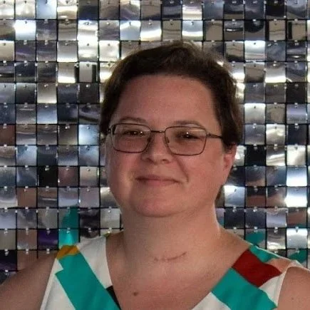 A woman with short dark hair and glasses, smiling, standing in front of a shiny, reflective, mosaic tile background.