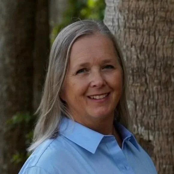 A woman with long gray hair smiling outdoors, standing in front of trees, wearing a light blue collared shirt.