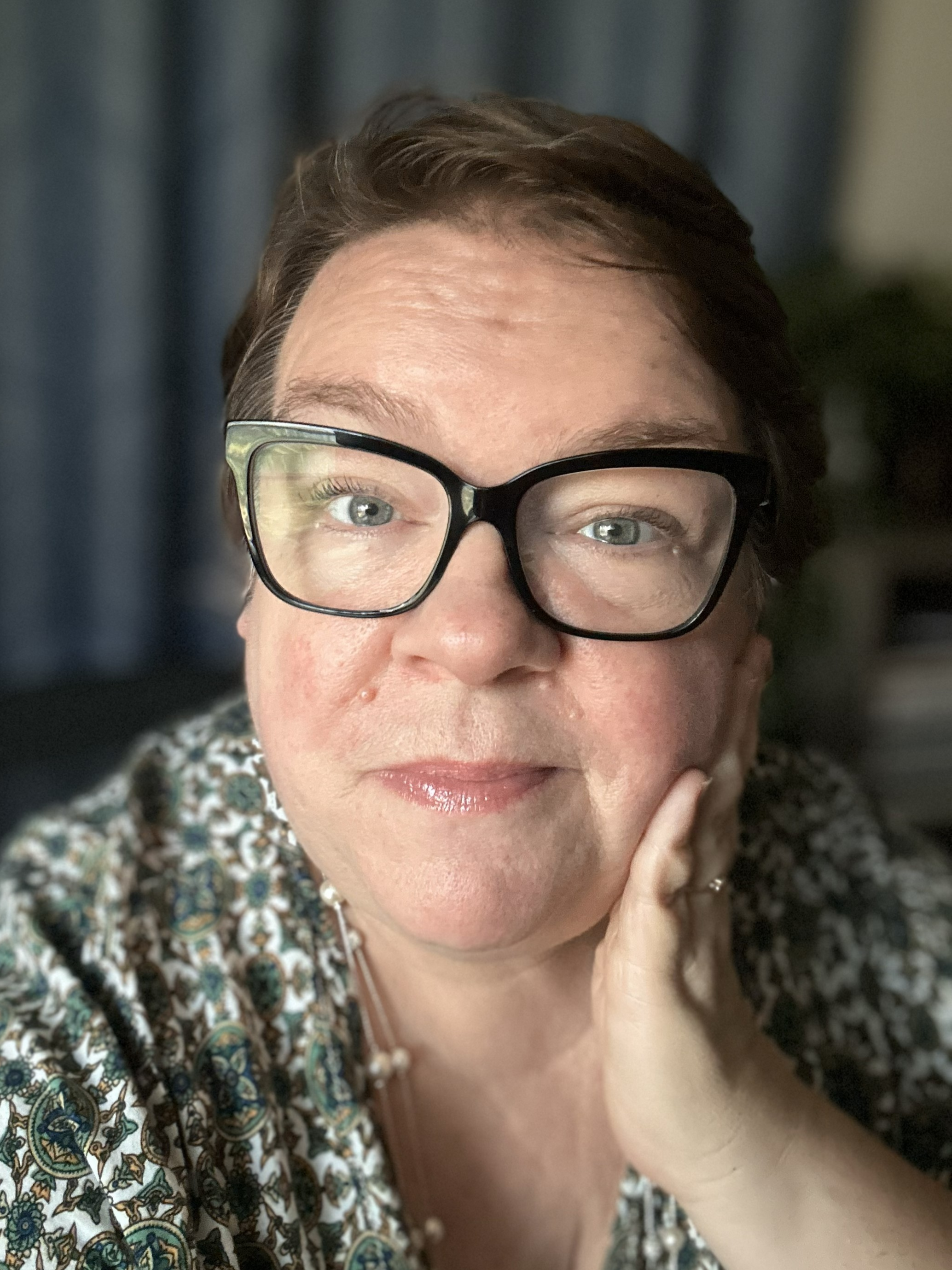 A close-up of a woman with short brown hair and glasses, wearing a patterned top, resting her chin on her hand.