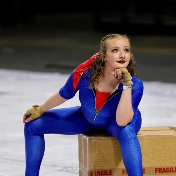 Woman dressed as a superhero in a blue suit with a red cape, sitting on a cardboard box labeled "Fragile," with a thoughtful expression and resting her chin on her hand.