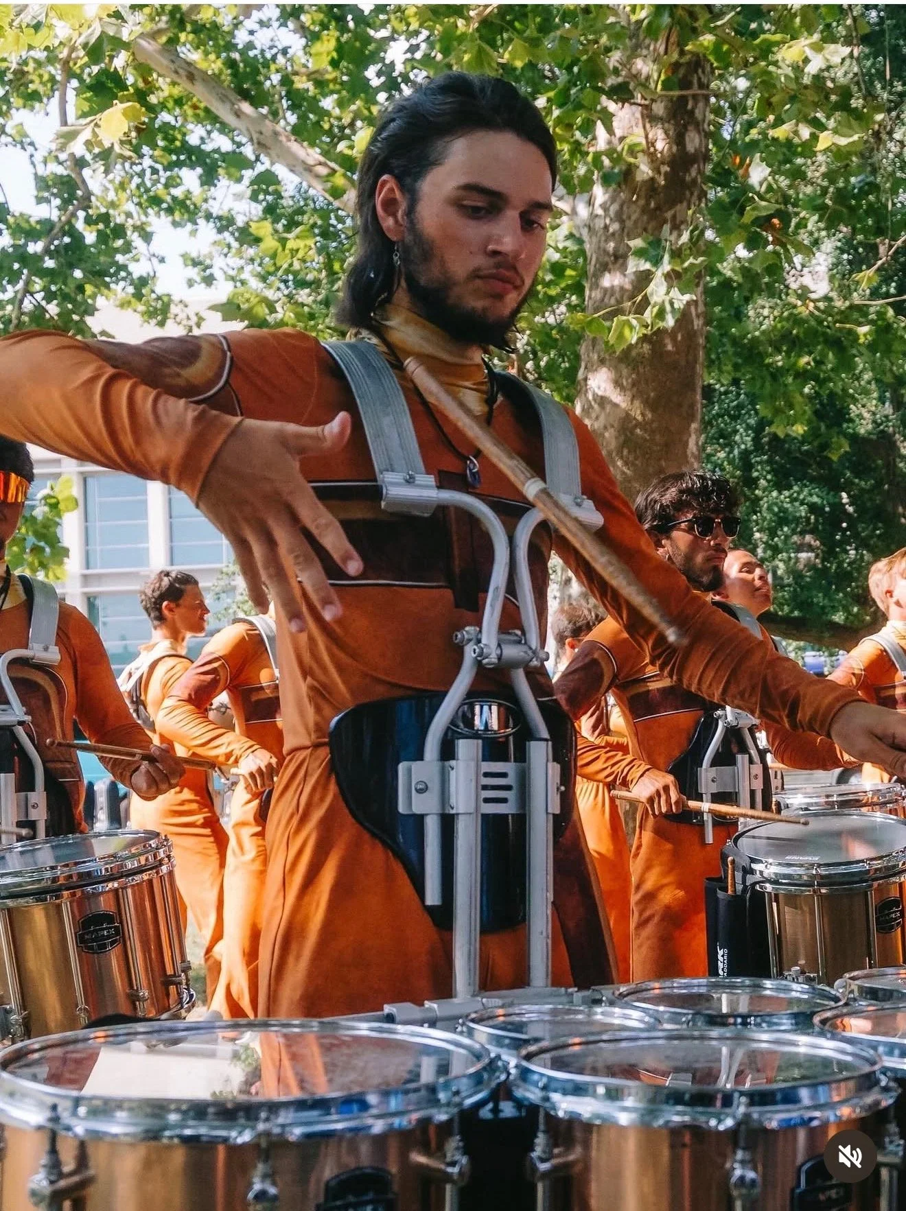 A group of musicians in orange outfits playing drums outdoors among green trees.