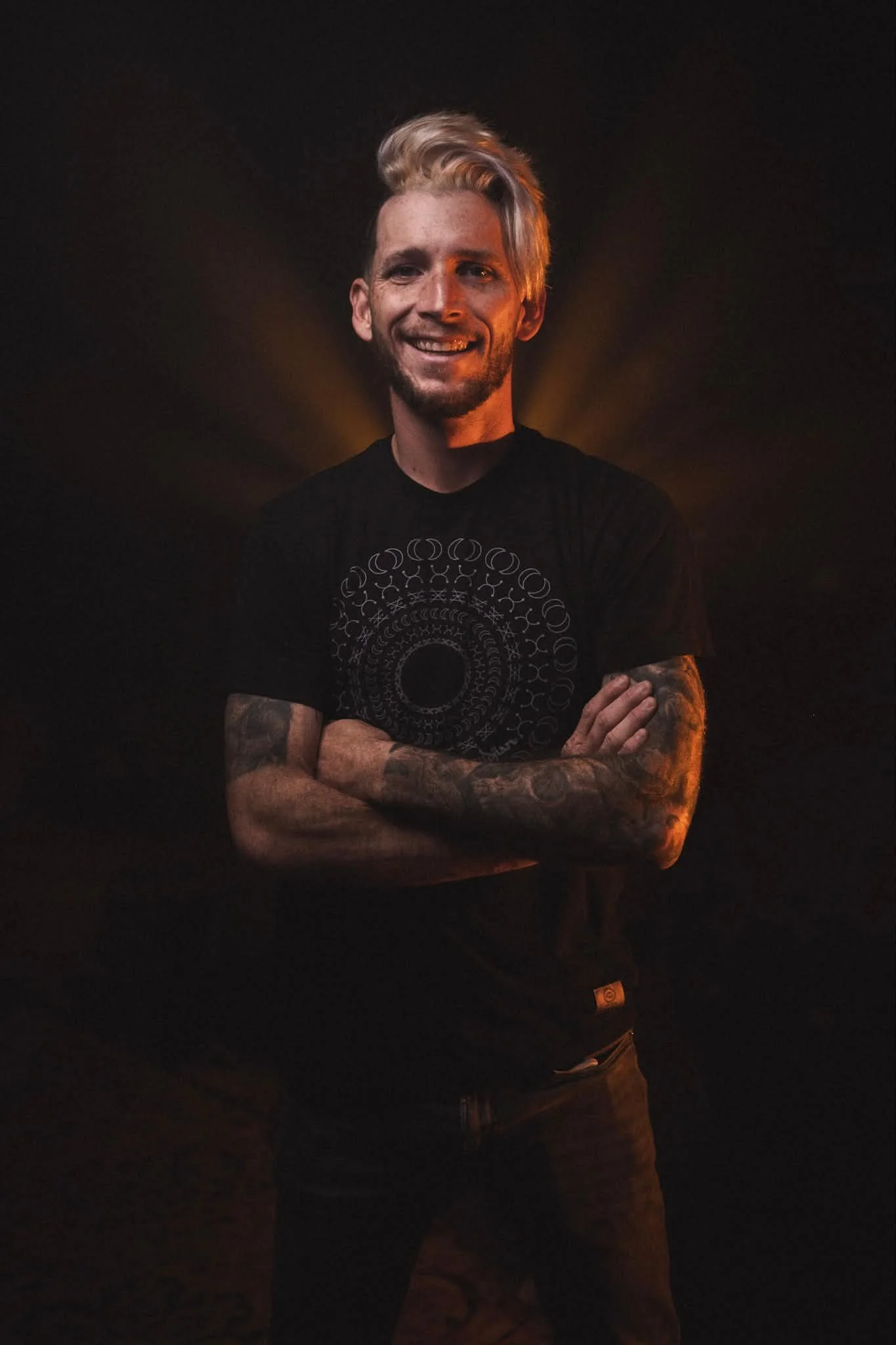 A man with blonde hair, tattoos on his arms, smiling and crossing his arms, wearing a black t-shirt with a geometric pattern, standing against a dark background with warm lighting.