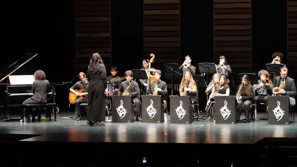 A school jazz band performing on stage with a pianist, guitarist, under the direction of a conductor, in a concert hall.