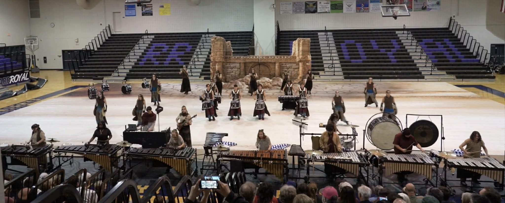 Performers in a gymnasium stage playing percussion instruments, with some dressed in costumes, and a backdrop resembling rocky ruins.