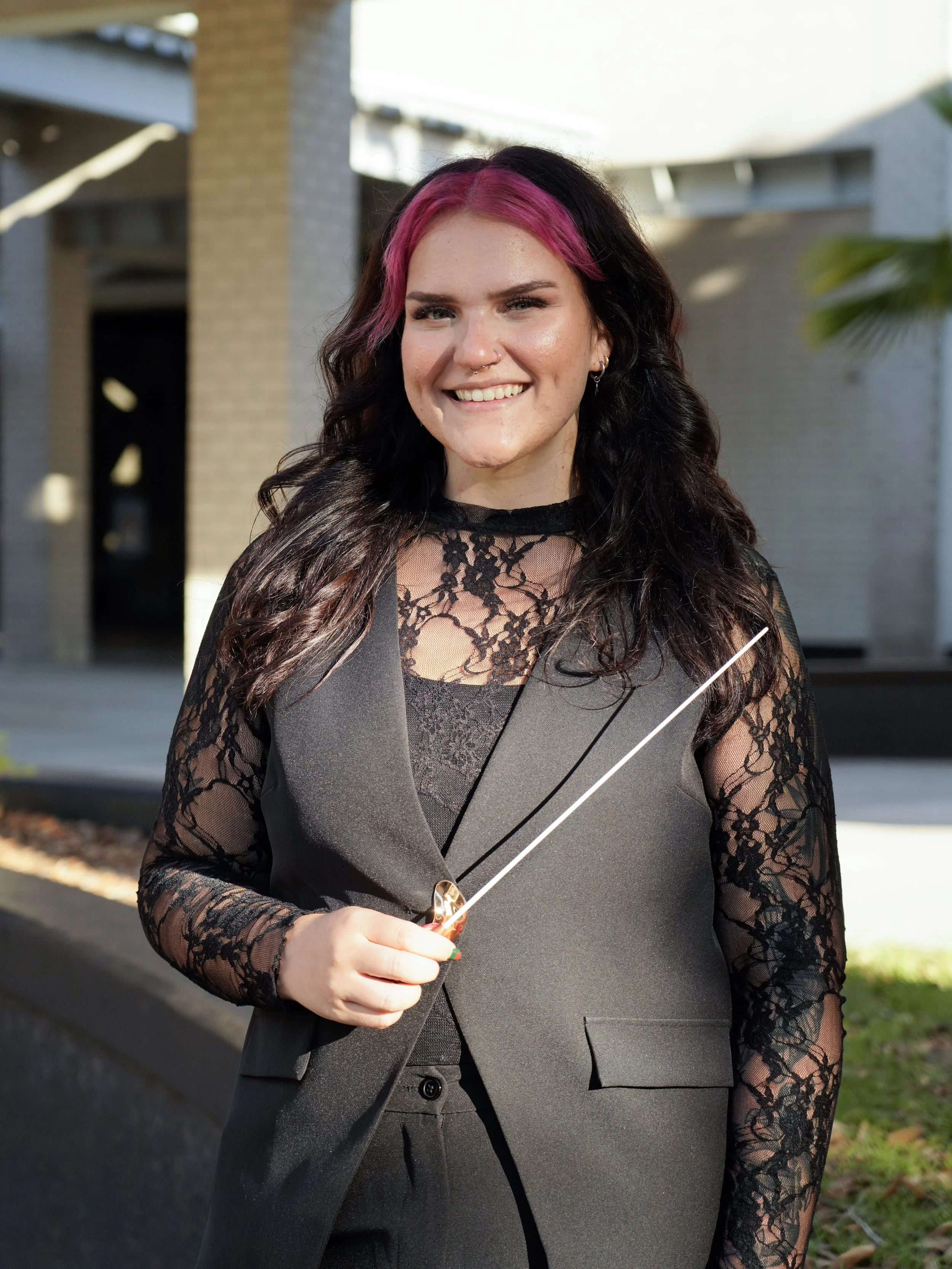 A woman with pink and black hair wearing a black lace top and gray vest, holding a drink with a straw, standing outdoors with modern building background.