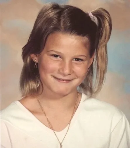 A young girl with light brown hair tied in a high ponytail, wearing a white shirt and gold jewelry, smiling at the camera.