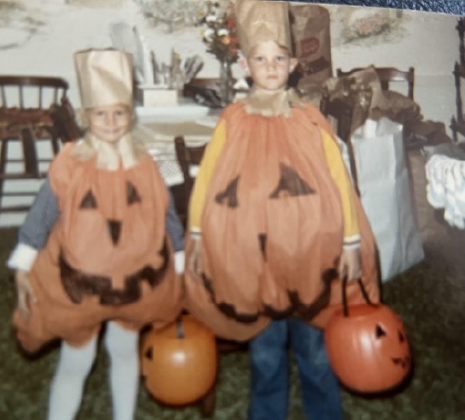 Two children dressed in Halloween costumes as pumpkins, holding pumpkin-shaped buckets for trick-or-treating, outdoors during daytime.