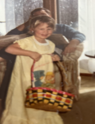 A young girl in a yellow dress holding a basket, standing next to an adult sitting on a couch in a cozy living room.