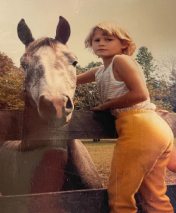 A young girl with short blonde hair stands next to a gray horse with a white face, touching its neck behind a wooden fence outdoors.