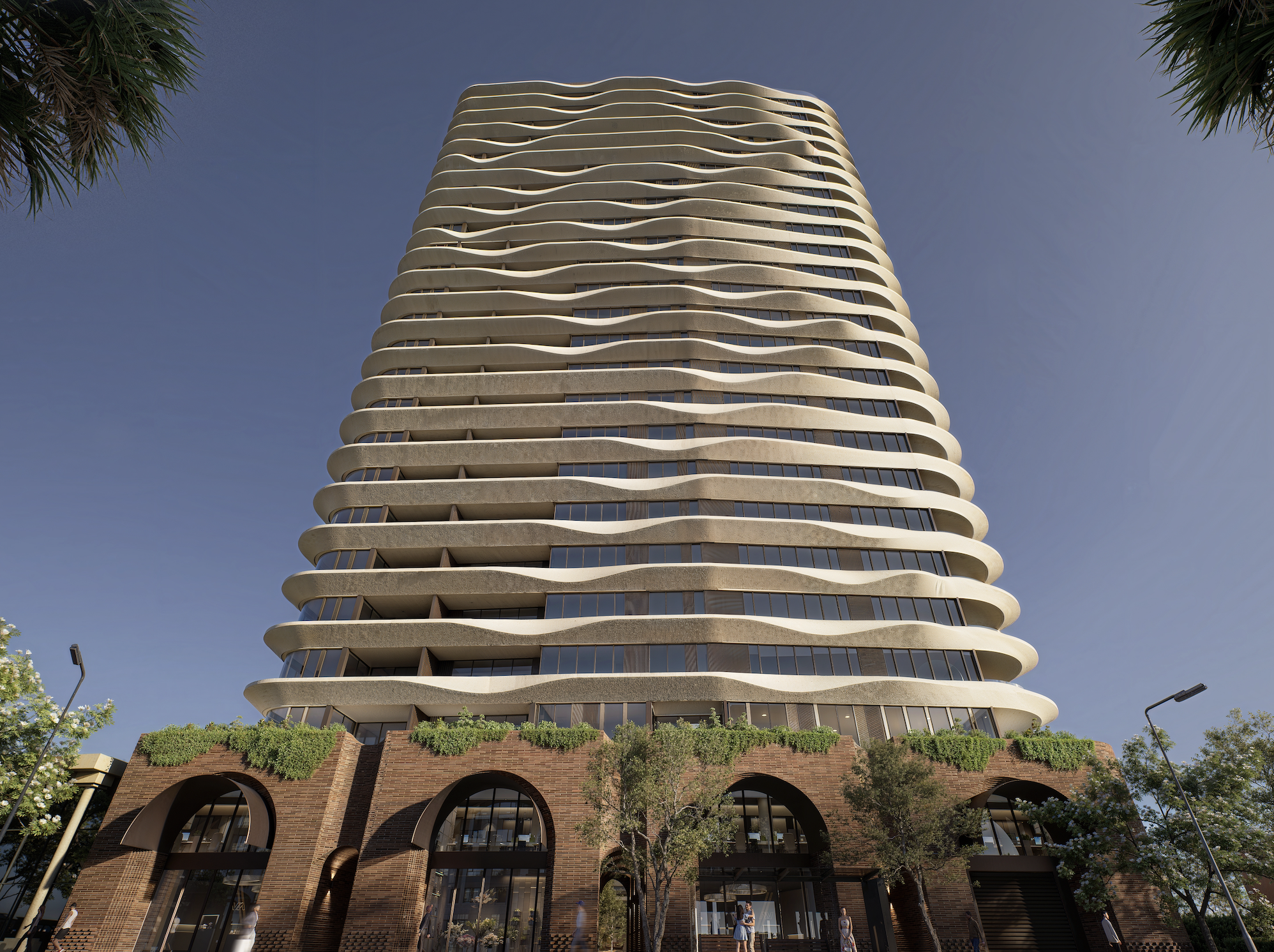 A modern multi-story building with a distinctive wavy architecture, featuring curved white balconies and glass windows, with trees and streetlights in the foreground and a clear blue sky above.