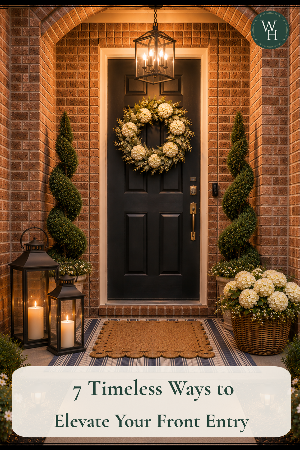 Timeless brick front entry with a black door, cream floral wreath, spiral topiaries, candle lanterns, layered doormats, and warm exterior lighting.