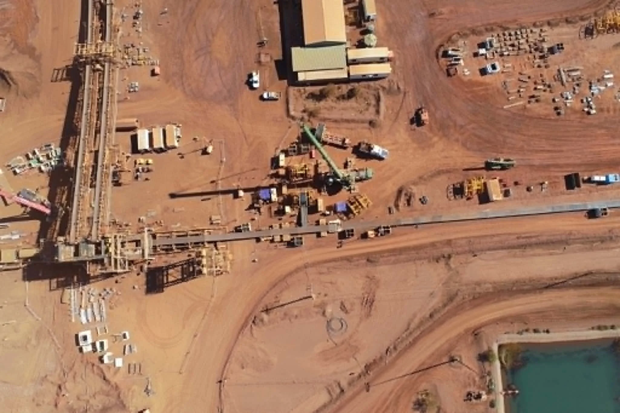 An aerial view of a construction site with an unfinished building, construction vehicles, and materials on reddish-brown terrain, alongside a small body of water and progression of roads.