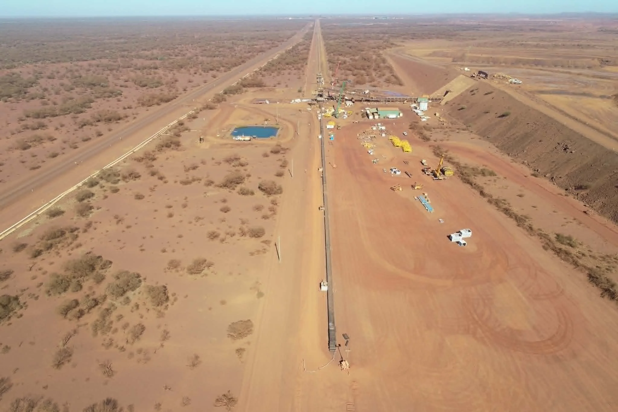 An aerial view of a construction site in a desert landscape, with equipment, vehicles, and a long pipeline under construction.