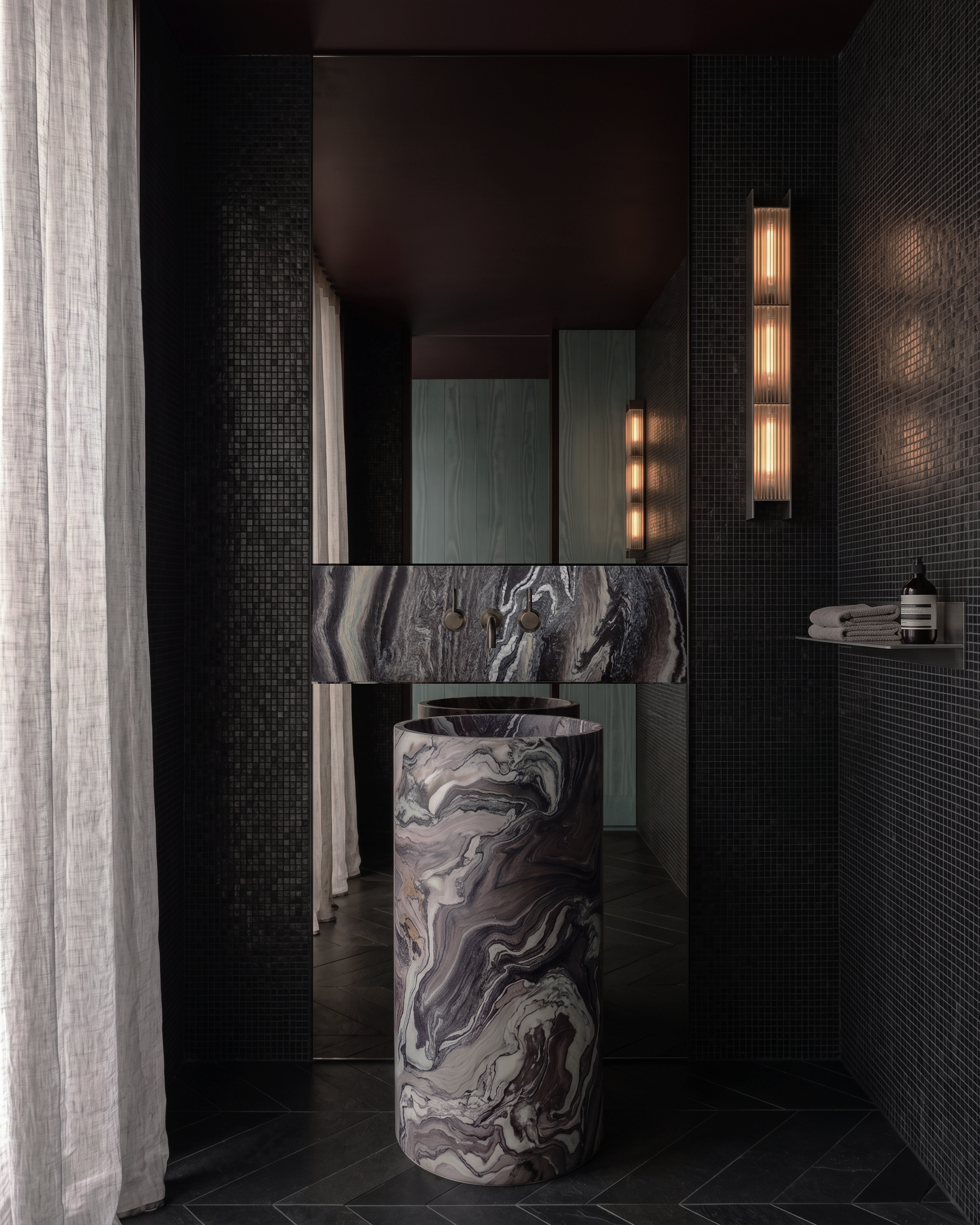 Modern bathroom with black tiled walls, a marble sink and matching cylindrical pedestal, a large mirror, and dim warm lighting.