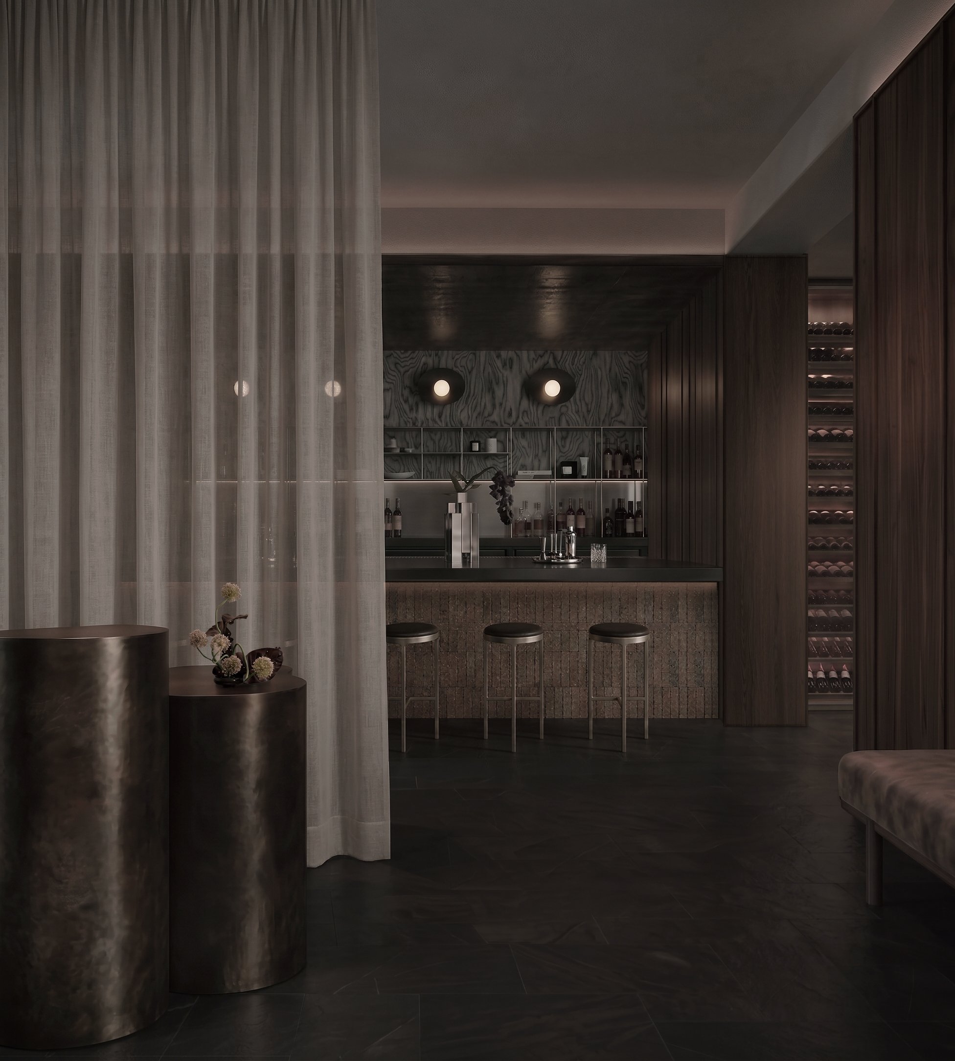 Interior view of a modern bar area with a dark color scheme. There are three metallic bar stools, a small vase with flowers on the counter, and a well-stocked shelf with bottles behind the bar. The space features textured wall paneling and warm lighting.