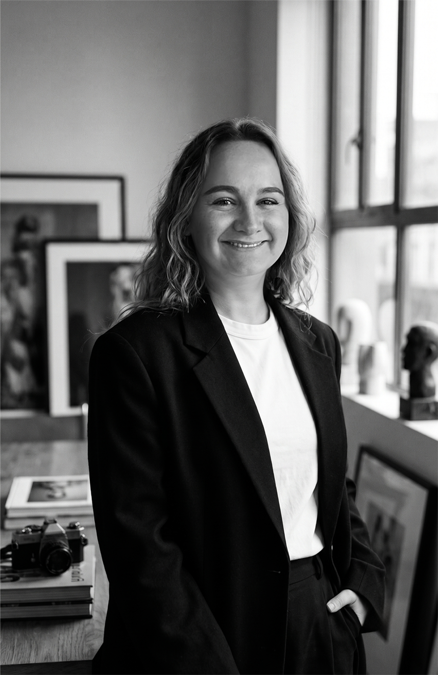 A smiling woman with wavy hair wearing a black blazer and white shirt standing in an art gallery or studio with framed artwork, sculptures, and a camera on a table behind her, near a window.