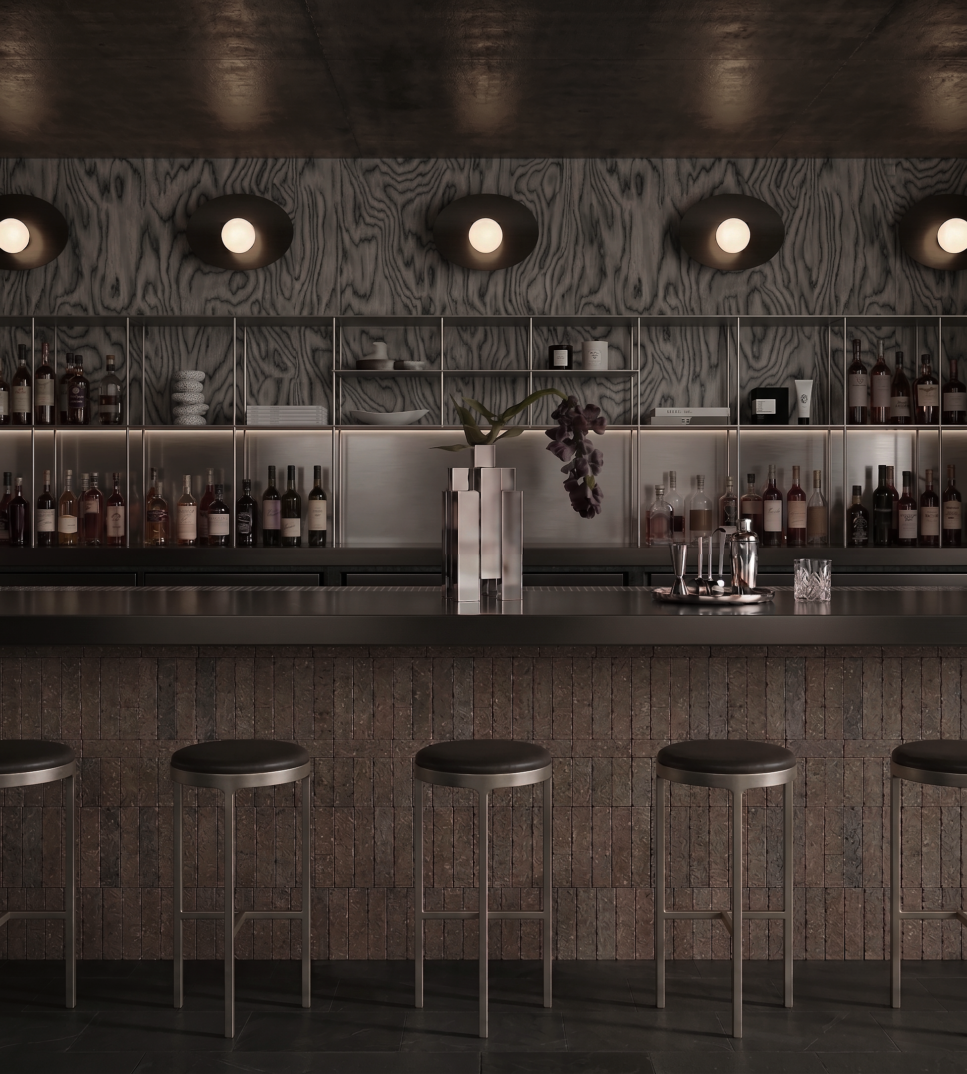 A modern bar counter with a dark tiled front, a sleek metal railing, and a marble countertop with bar tools and a flower arrangement, set against a textured wood and marble wall with five exposed lightbulbs on the wall.