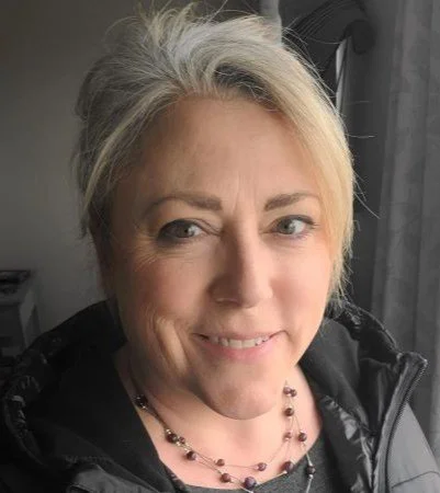A woman with blonde hair styled in a casual updo, smiling indoors near a window with curtains, wearing a black jacket and a beaded necklace.