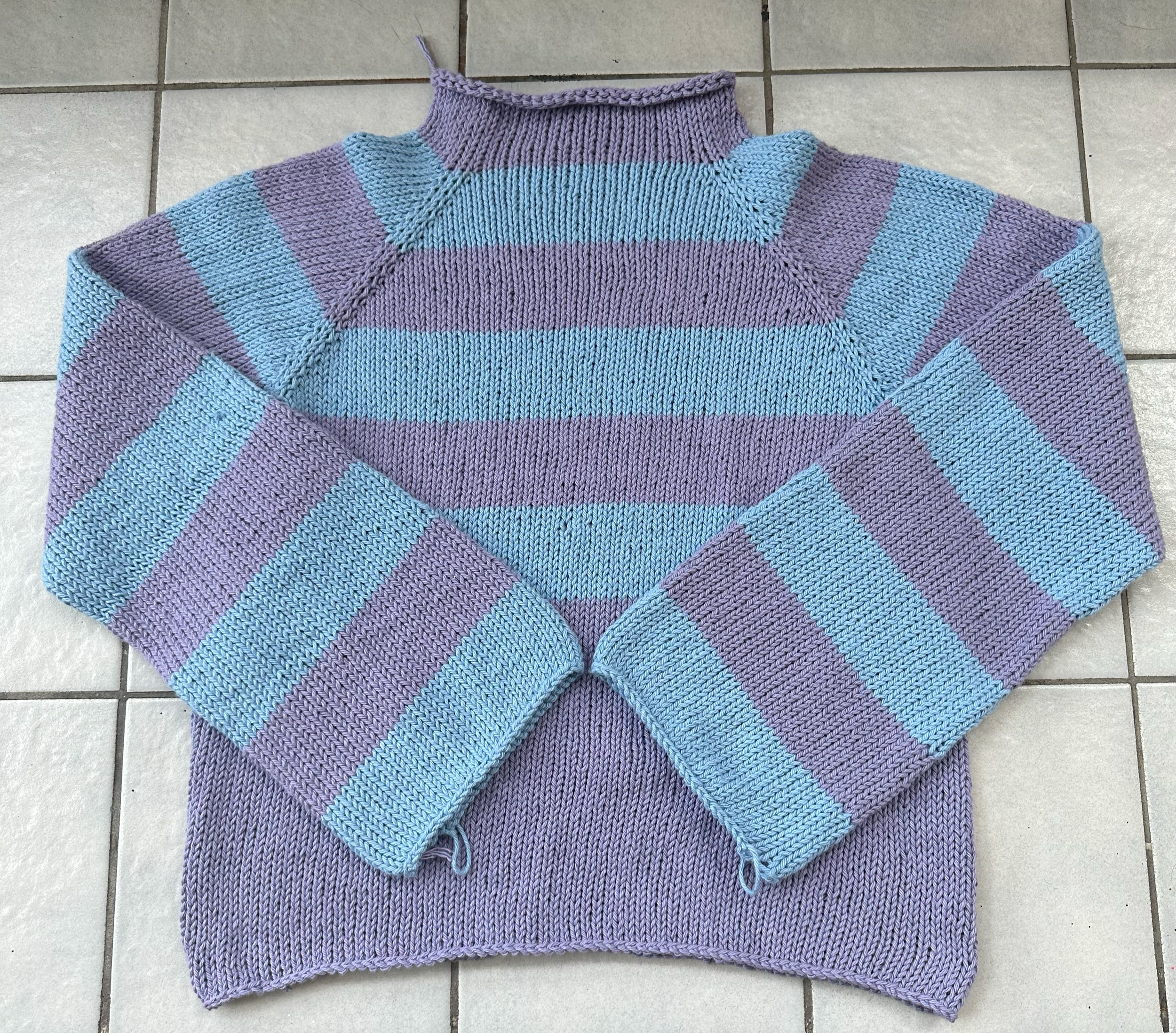 Hand-knitted striped sweater in shades of purple and blue, laying on a tiled floor.