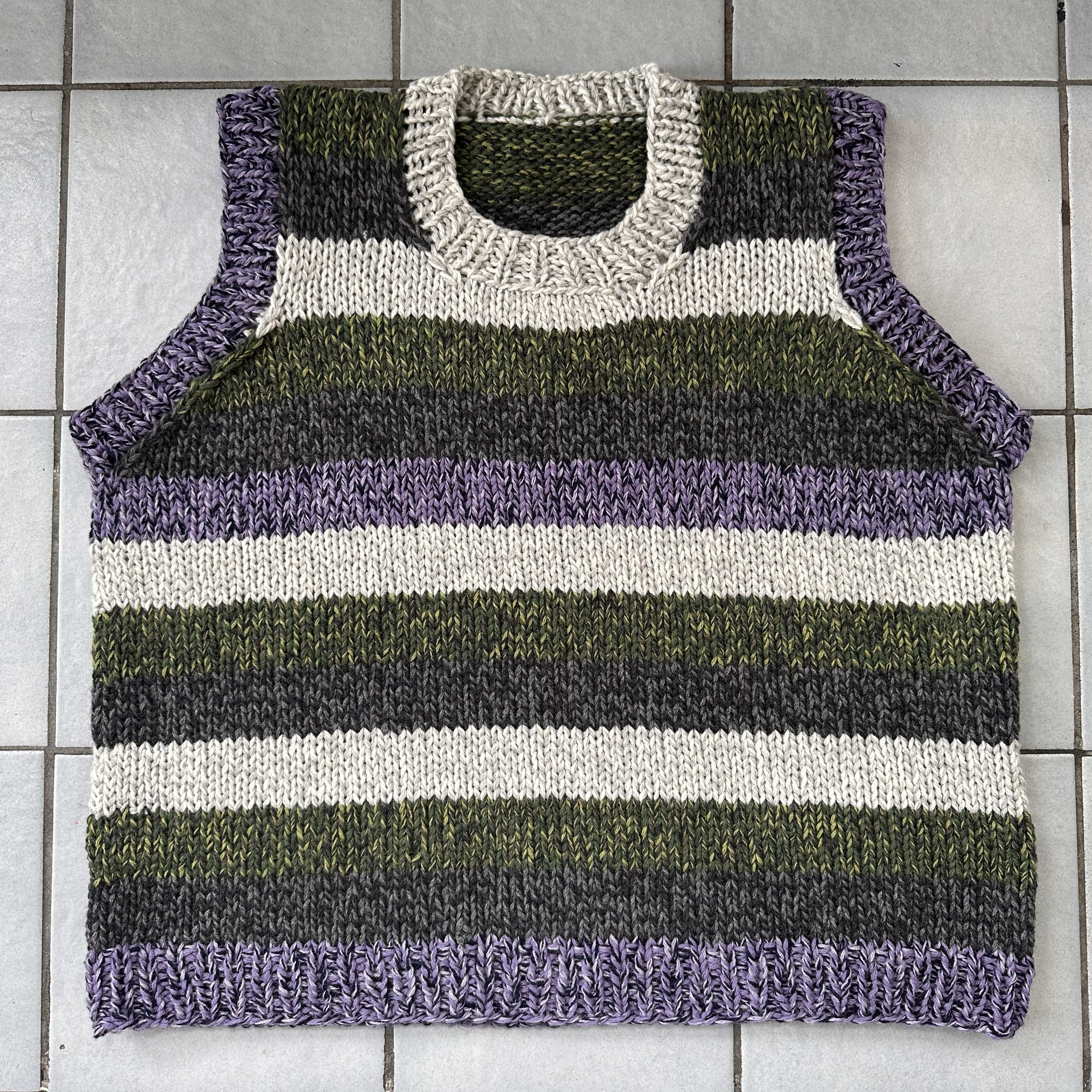 Knitted sweater with purple, green, black, and white stripes laid flat on a tiled floor.