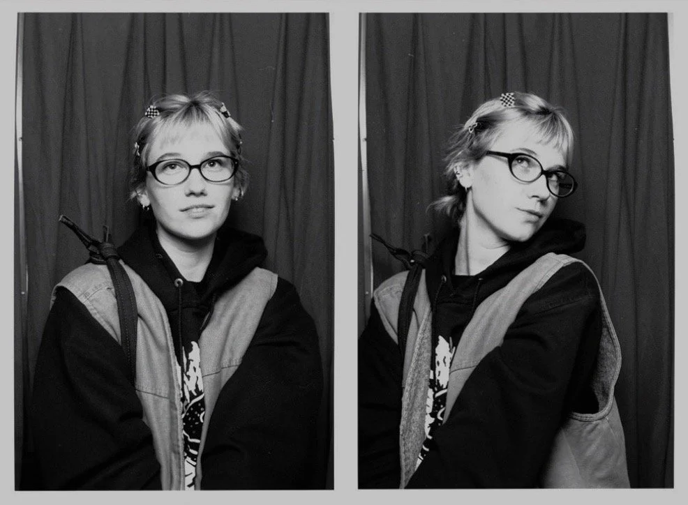 Black and white photo of a young woman with short hair, wearing glasses, a hoodie, and a vest. She is posing with two different expressions in front of a curtain backdrop.