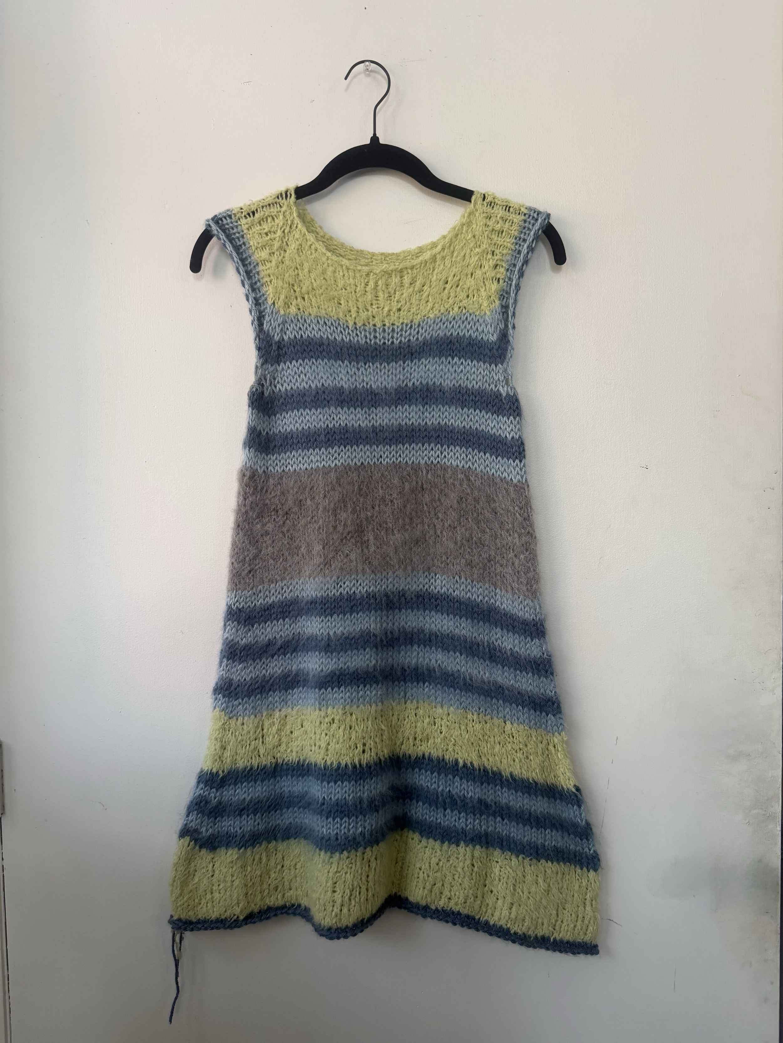 Handmade knitted dress with horizontal color stripes in yellow, gray, blue, and brown, hanging on a black hanger on a white wall.