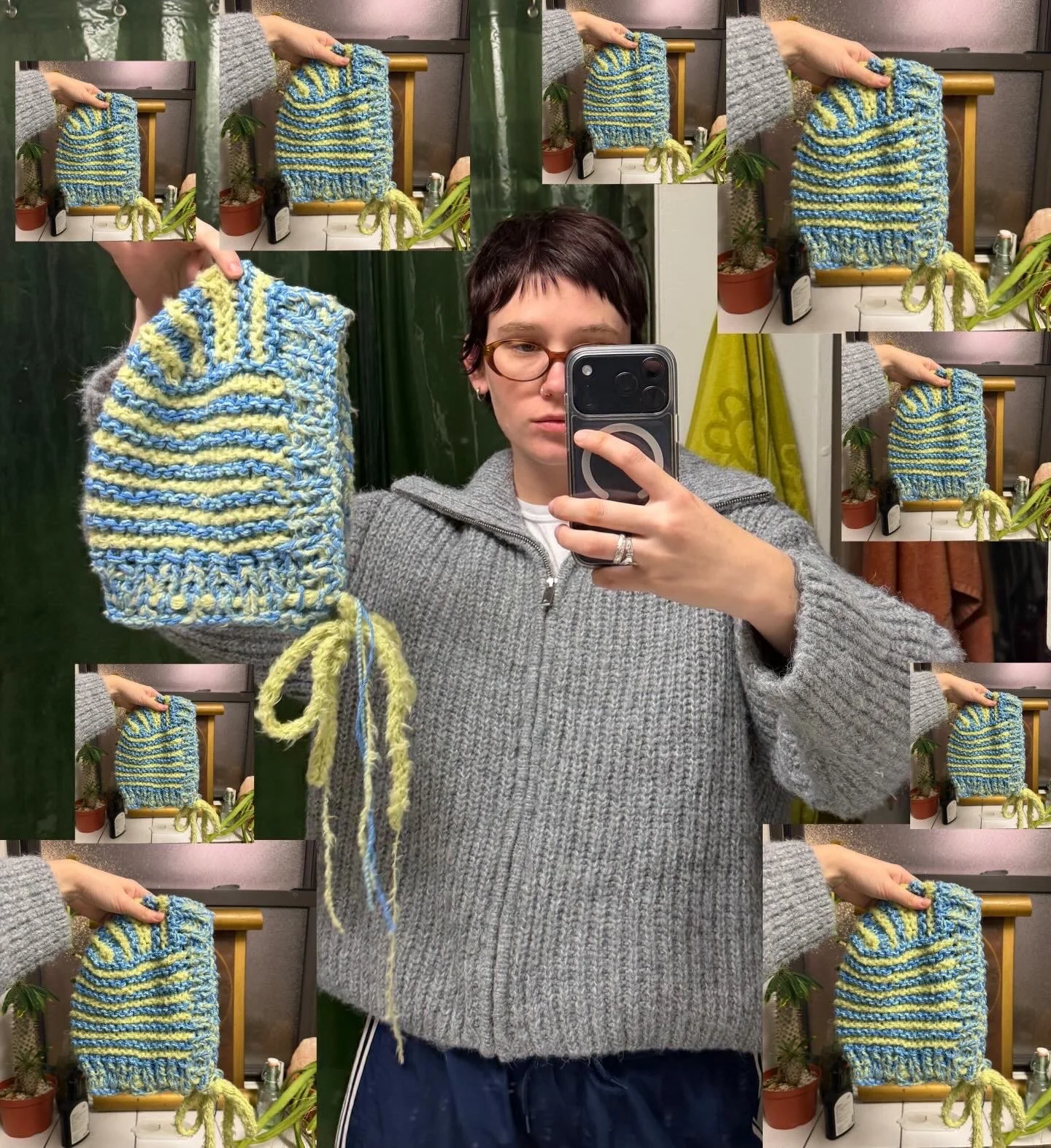 A woman taking a mirror selfie in a room with green plants and a yellow curtain, holding a small colorful striped knitted bag or sock near the mirror, wearing glasses and a gray knitted sweater.