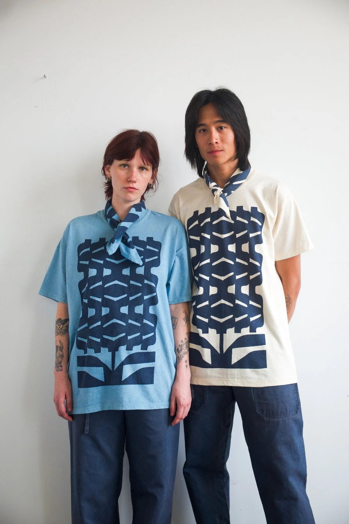 Two people standing against a plain white wall, wearing matching graphic T-shirts and bandanas around their necks. The person on the left has short red hair, and the person on the right has long black hair.