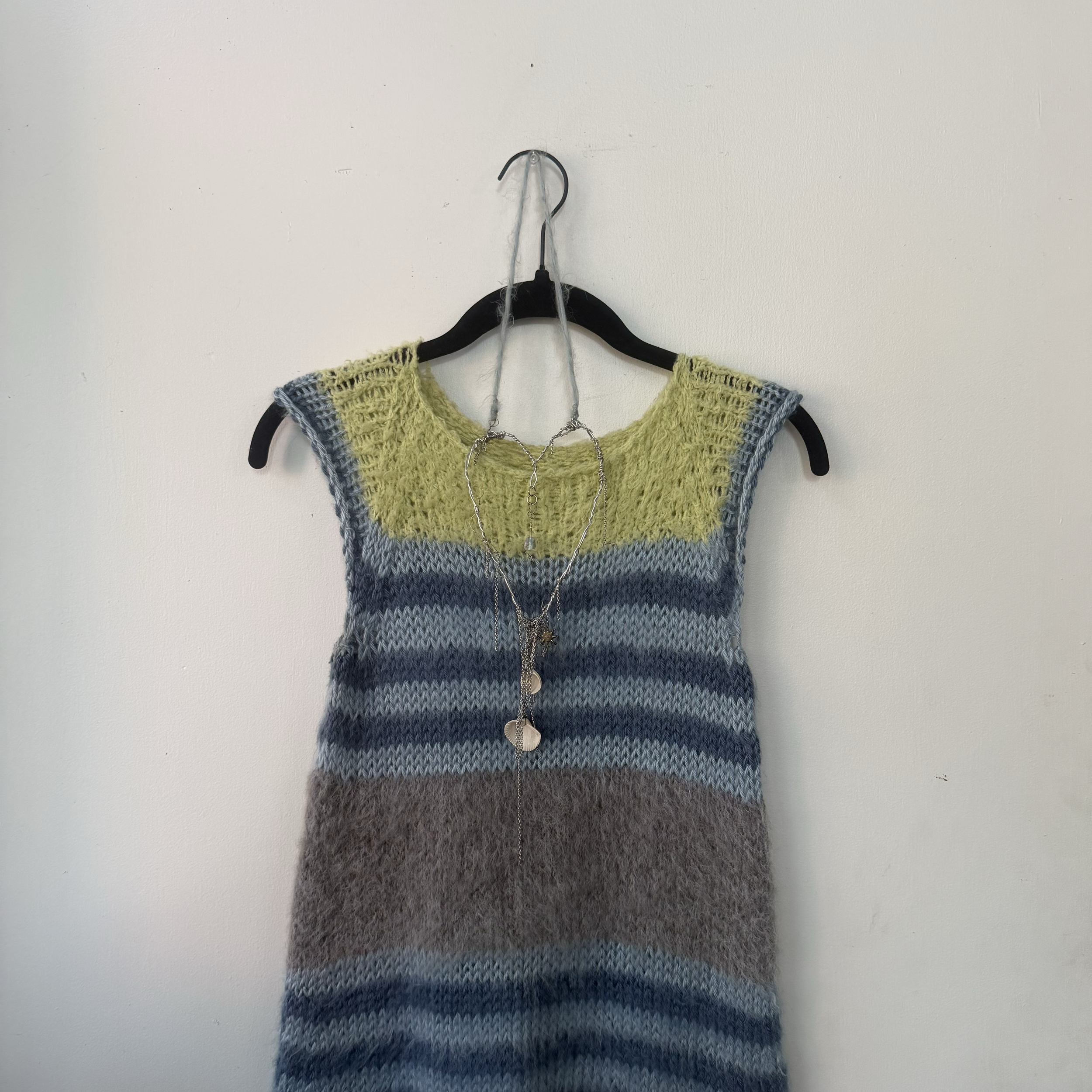 A multicolored, knitted sleeveless top hanging on a black hanger, which is suspended from a nail on a plain white wall.