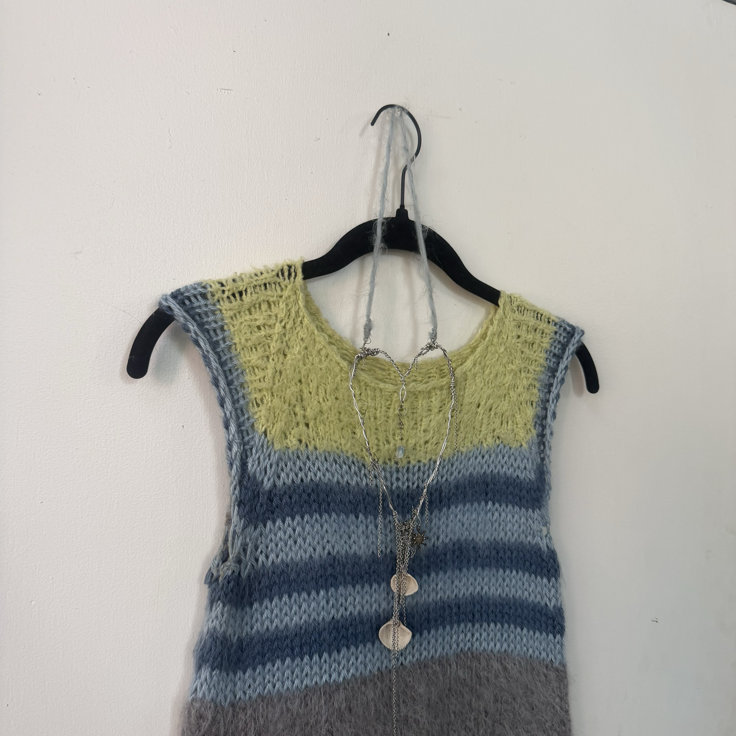 A black clothing hanger with a knitted yellow and gray sweater hanging on it. The hanger is on a metal hook attached to a plain white wall. Several silver jewelry pieces, including necklaces with seashell and starfish charms, are draped over the swea