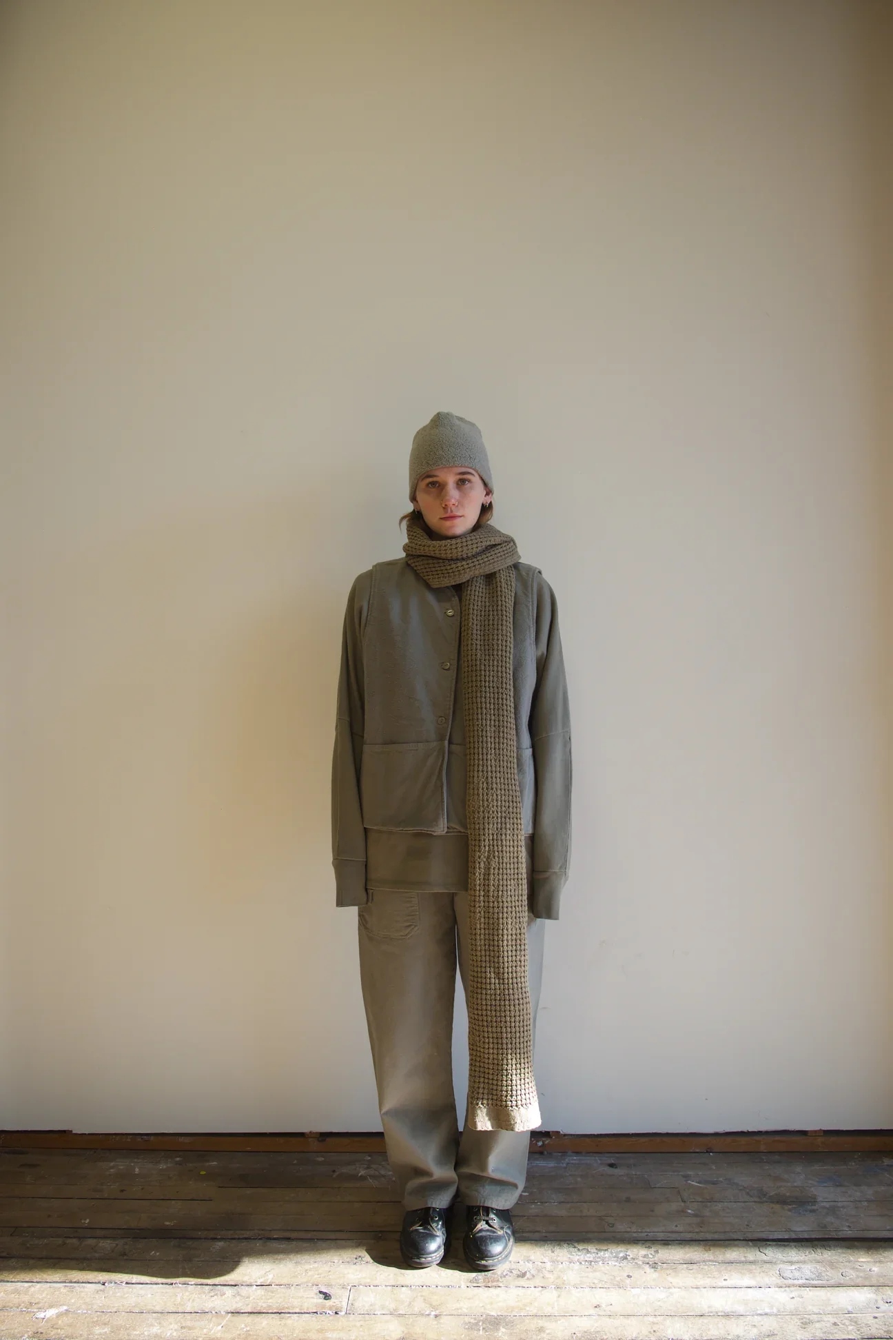 A person standing against a plain light-colored wall, wearing a gray hat, a matching gray jacket and pants, black shoes, and a long checkered scarf.