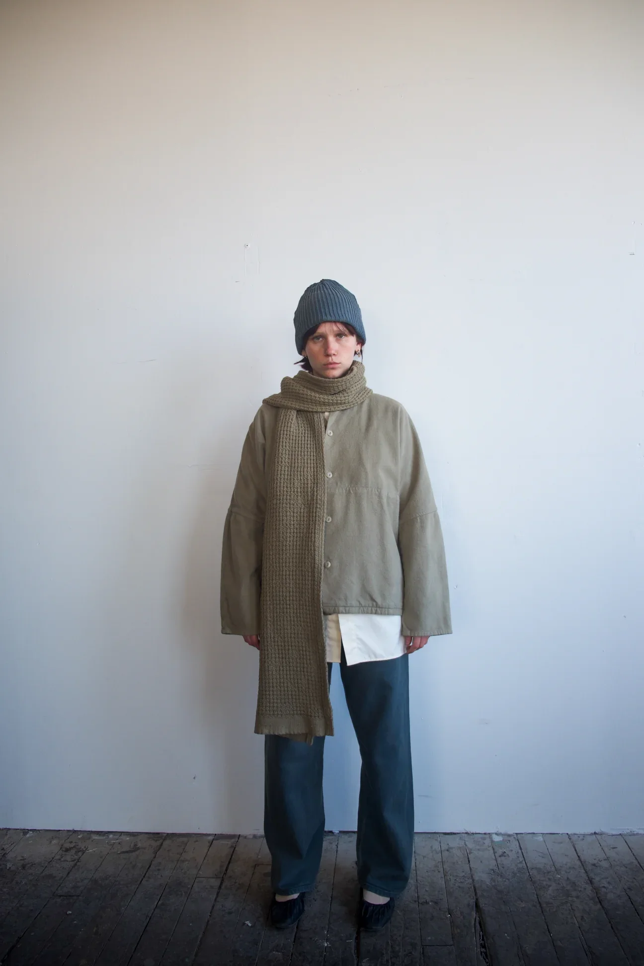 Person standing against a plain white wall, wearing a blue knit beanie, beige loose shirt, a long brown knitted scarf, dark blue wide-leg jeans, and dark shoes.