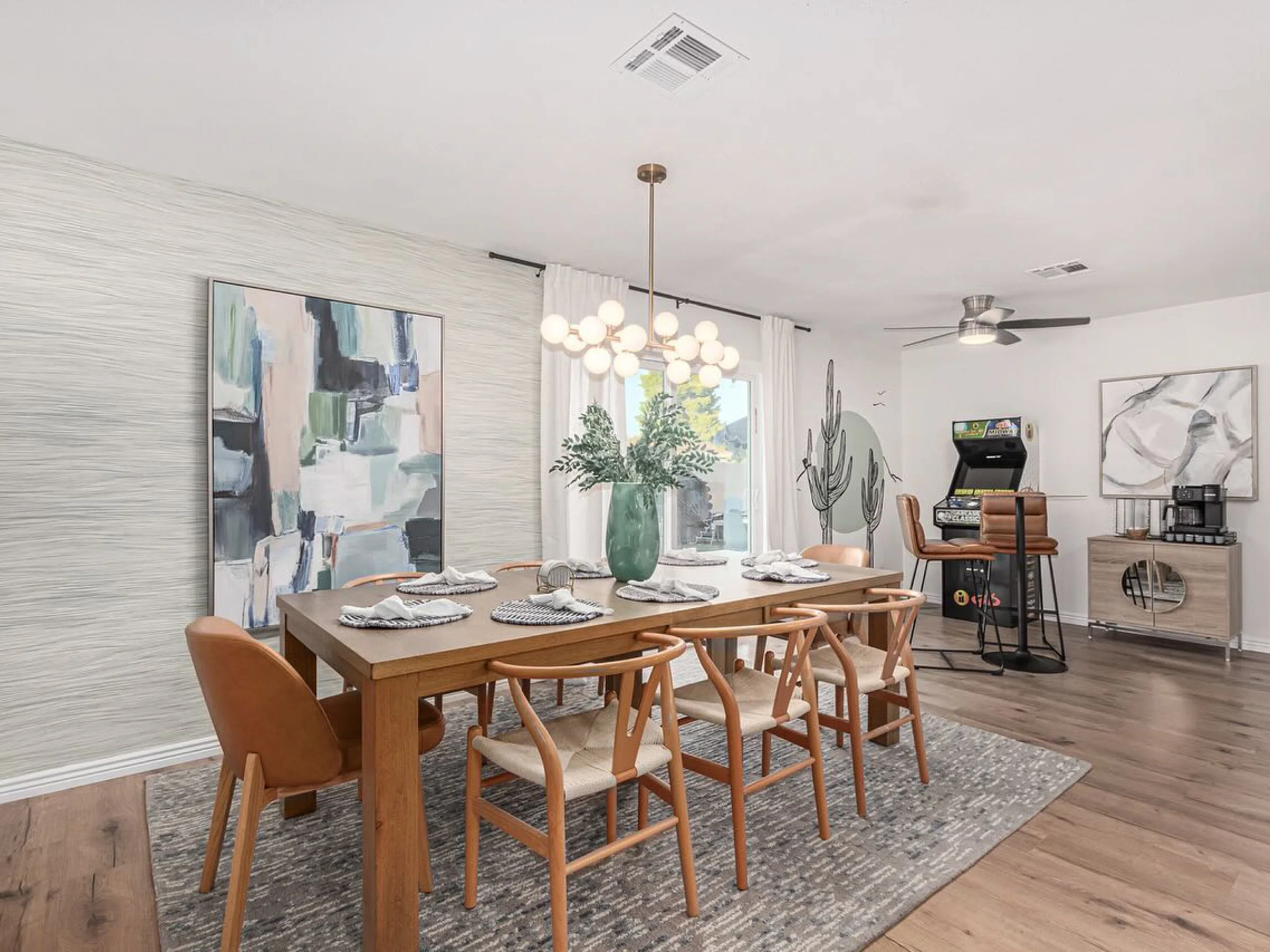 An interior dining area with a wooden table set with plates and napkins, surrounded by wooden chairs, a large window with white curtains, a modern chandelier, wall art, and a living space with a ceiling fan and artwork.