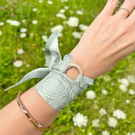 A person's wrist decorated with a silver ring, a silver bracelet, and a gray bandana tied around the wrist with a circular ring. The background features small white flowers and green grass.