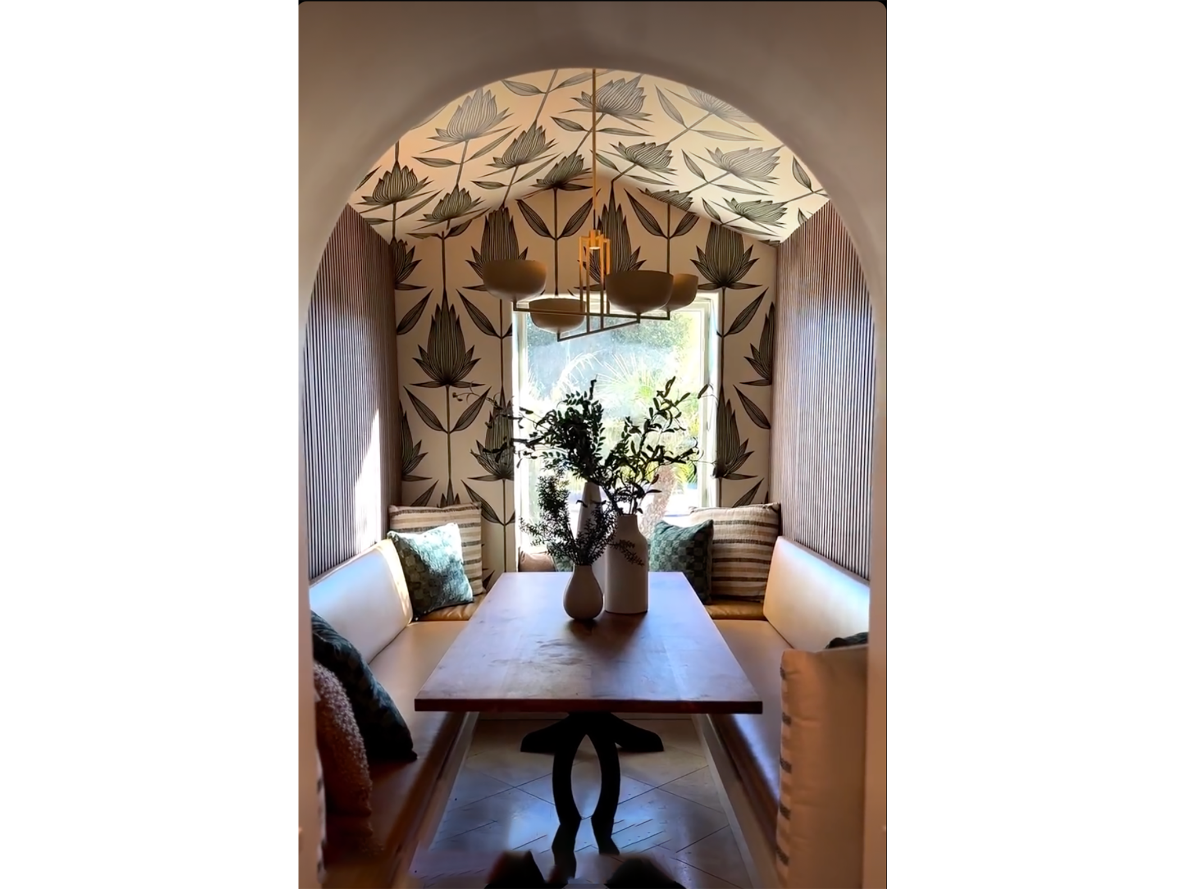 A dining nook with a wooden table, beige cushioned seating, and pillows, surrounded by textured wall paneling and wallpaper with large floral patterns. A window lets in natural light, and a decorative chandelier hangs above the table with a vase of plants in the center.