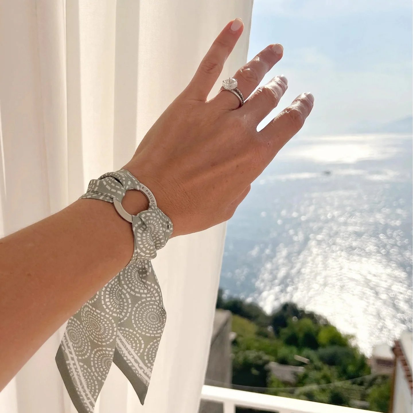 Close-up of a woman's left hand with an engagement ring, wristwatch, and bracelet, reaching towards a bright ocean view with sunlight reflecting on the water.