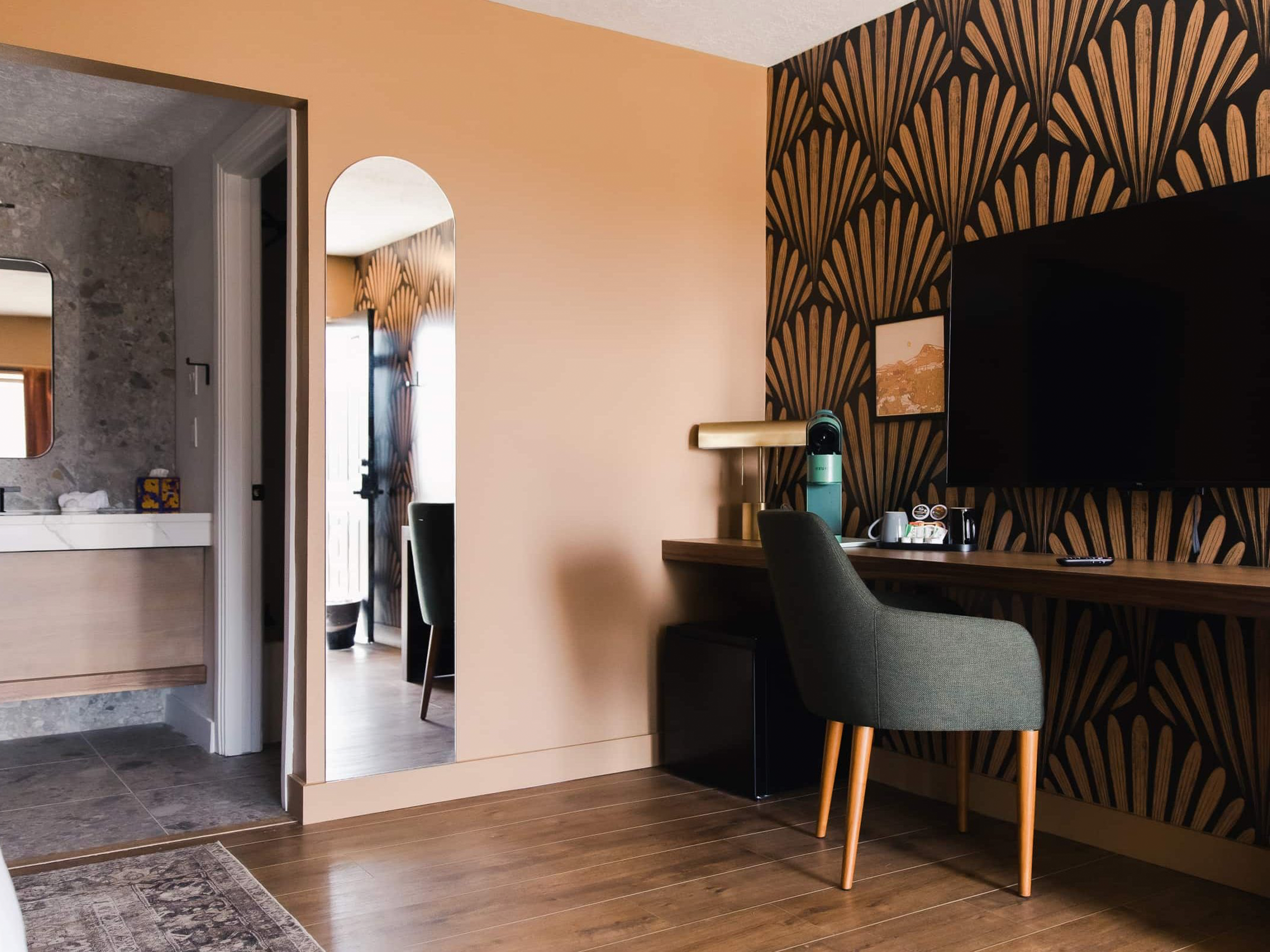 A section of a modern interior room featuring a black and gold geometric patterned wallpaper, a wooden desk with a grey upholstered chair, a wall-mounted TV, and a gold lamp. A doorway shows part of an adjacent room with a mirror and a pizza box.