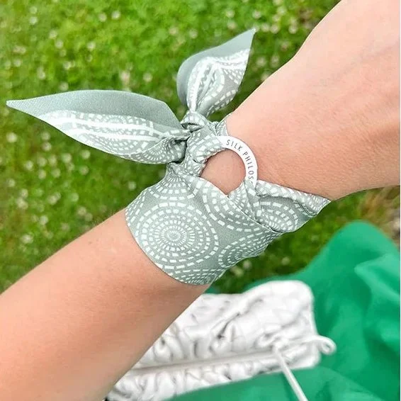 A person's wrist with a light gray bandana tied around it, with a small fabric hole, and a white circular tag that reads 'SILK PILLOW.' The background shows green grass and part of a white item, possibly clothing or fabric.