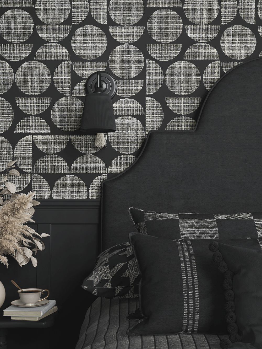 Close-up of a bedroom corner with a black textured headboard, patterned pillows, a wall-mounted black lamp, a patterned wallpaper background, and a side table holding a cup, saucer, and books.